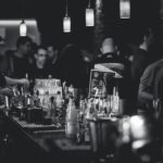 Black and white photo capturing the lively atmosphere in a bar with people socializing and a busy bartender serving drinks.