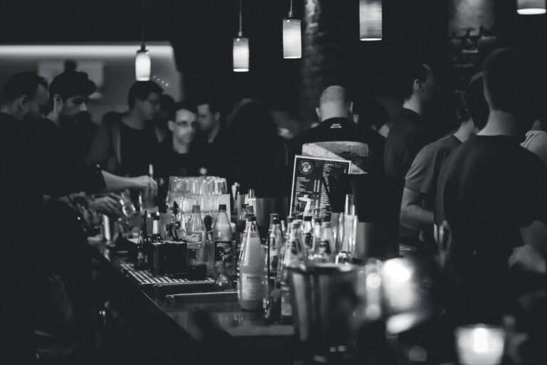 Black and white photo capturing the lively atmosphere in a bar with people socializing and a busy bartender serving drinks.